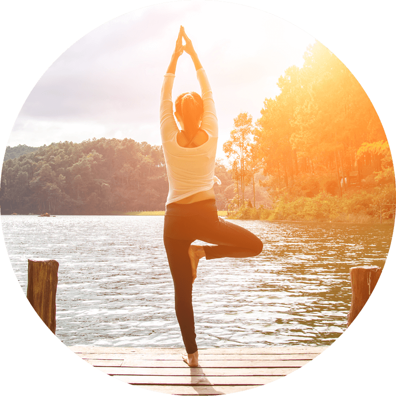 Eversense user practicing yoga tree pose on a wooden dock, surrounded by a tranquil lake and forested hills, with sunlight.