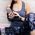 Woman wearing gym clothes uses smartphone, sitting on floor beside a pink water bottle, brick wall in background.