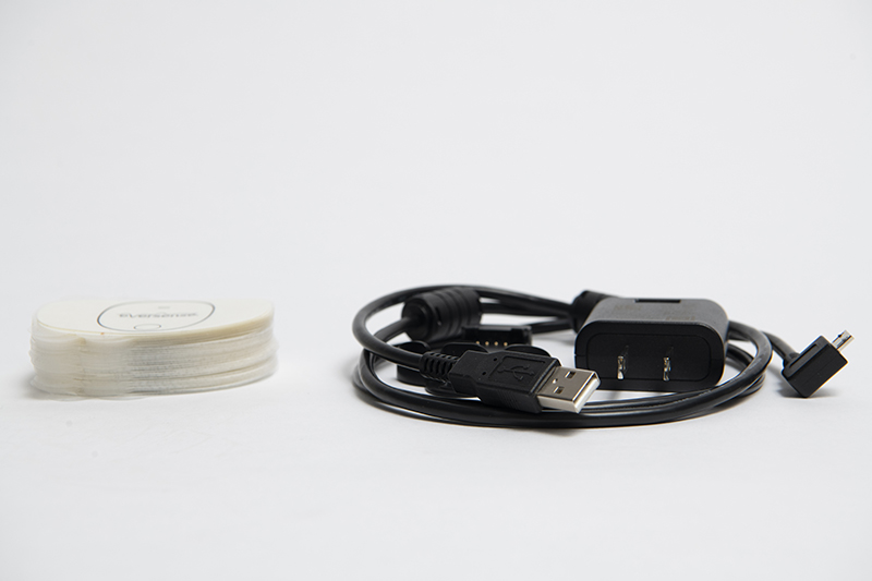 A USB charging cable with adapter lies coiled on a flat surface next to transmitter adhesives